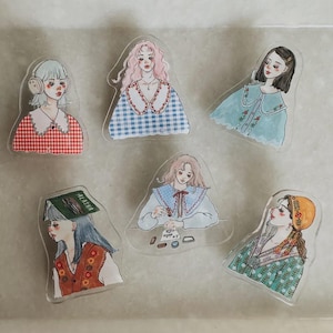 May include: Six clear stickers with illustrations of women in diverse outfits and hairstyles. The stickers display a variety of colours, including red, blue, and green, with detailed artwork. These stickers are suitable for decorating journals, laptops, or other items.
