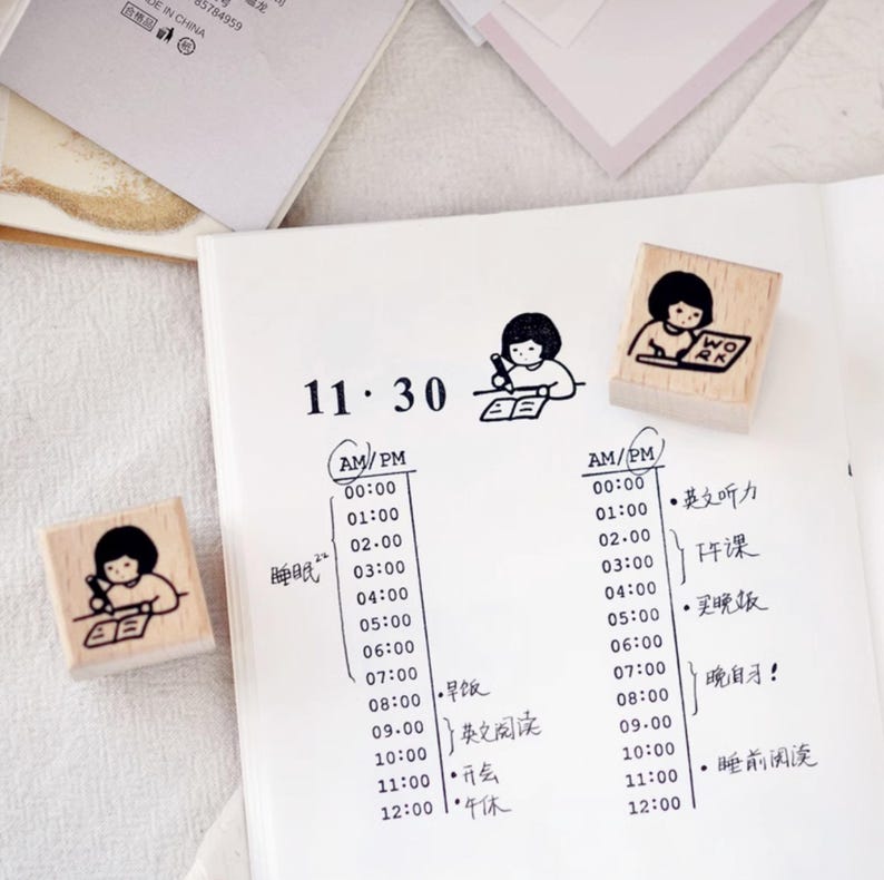 Puede incluir: Sellos de madera con un dise&ntilde;o de ni&ntilde;a de dibujos animados, ideales para hacer un diario o planificar. Los sellos muestran a la ni&ntilde;a escribiendo y la palabra "WORK". La imagen tambi&eacute;n muestra una p&aacute;gina de planificador con horarios escritos a mano y caracteres chinos.