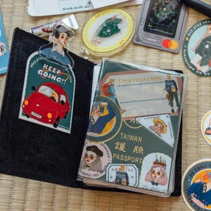 May include: A passport cover with a green background and a variety of stickers. The stickers feature illustrations of people, cars, and travel destinations. The cover has the text "Taiwan 護照 Passport" printed on it.