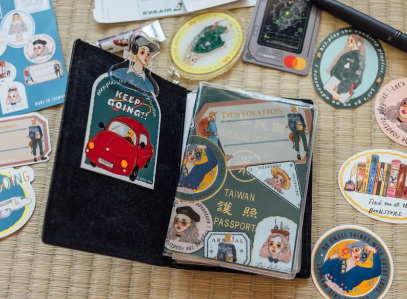 May include: A passport cover with a green background and a variety of stickers. The stickers feature illustrations of people, cars, and travel destinations. The cover has the text "Taiwan 護照 Passport" printed on it.
