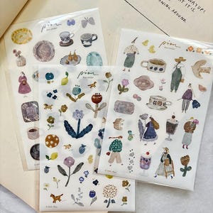 May include: Three sticker sheets featuring watercolor illustrations of teacups, plates, flowers, and figures. The stickers have a whimsical, artistic style with soft colors and the word "pion" printed on them. The background includes handwritten text.