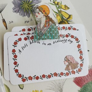 May include: Three white rectangular cards with a floral border and the text "A soft bloom in an ordinary day." A clear bookmark features a woman in a headscarf. Botanical illustrations are in the background.