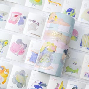 May include: A cylindrical 2027 washi tape calendar container featuring pastel watercolor illustrations of figures in various poses. The container is placed atop a matching accordion-style paper calendar sheet displaying dates and colorful artistic scenes.