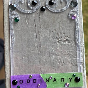 Puede incluir: Un marco de plástico transparente con un diseño de purpurina plateada y piedras de imitación. El marco tiene un fondo morado y verde con la palabra "ODDINARY" escrita en letras negras.