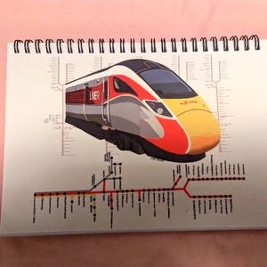 Può includere: Quaderno a spirale con un'illustrazione dettagliata di un treno ad alta velocità. Il treno presenta accenti rossi, arancioni e gialli ed è etichettato "LNER" e "AZUMA". Sotto il treno c'è una mappa stilizzata del percorso.