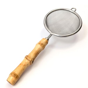 May include: A stainless steel tea strainer with a bamboo handle. The strainer has a fine mesh and a circular shape. The handle is made of light brown bamboo with a natural grain. The strainer is designed for steeping loose leaf tea.