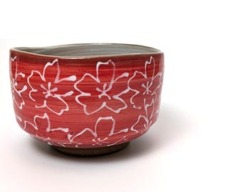 May include: A red ceramic bowl with a white floral pattern. The bowl has a slightly irregular shape and a gray interior. The base is a darker brown color. The floral design is hand-painted.