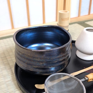 May include: A dark blue and brown ceramic tea bowl with a textured exterior. The bowl is square-shaped with rounded corners and sits on a black tray. Other tea ceremony tools are visible in the background.