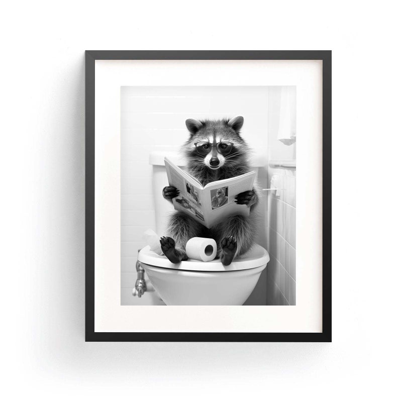 May include: A black and white framed print featuring a raccoon wearing round glasses while sitting on a white toilet and reading a newspaper. A roll of toilet paper sits on the seat next to the raccoon, all enclosed in a simple black frame.