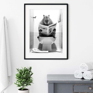 May include: A black and white photograph of a capybara sitting on a toilet and reading a newspaper. The capybara is in a bathroom setting with white tiles and a towel hanging on the wall. The photograph is framed in a black frame.
