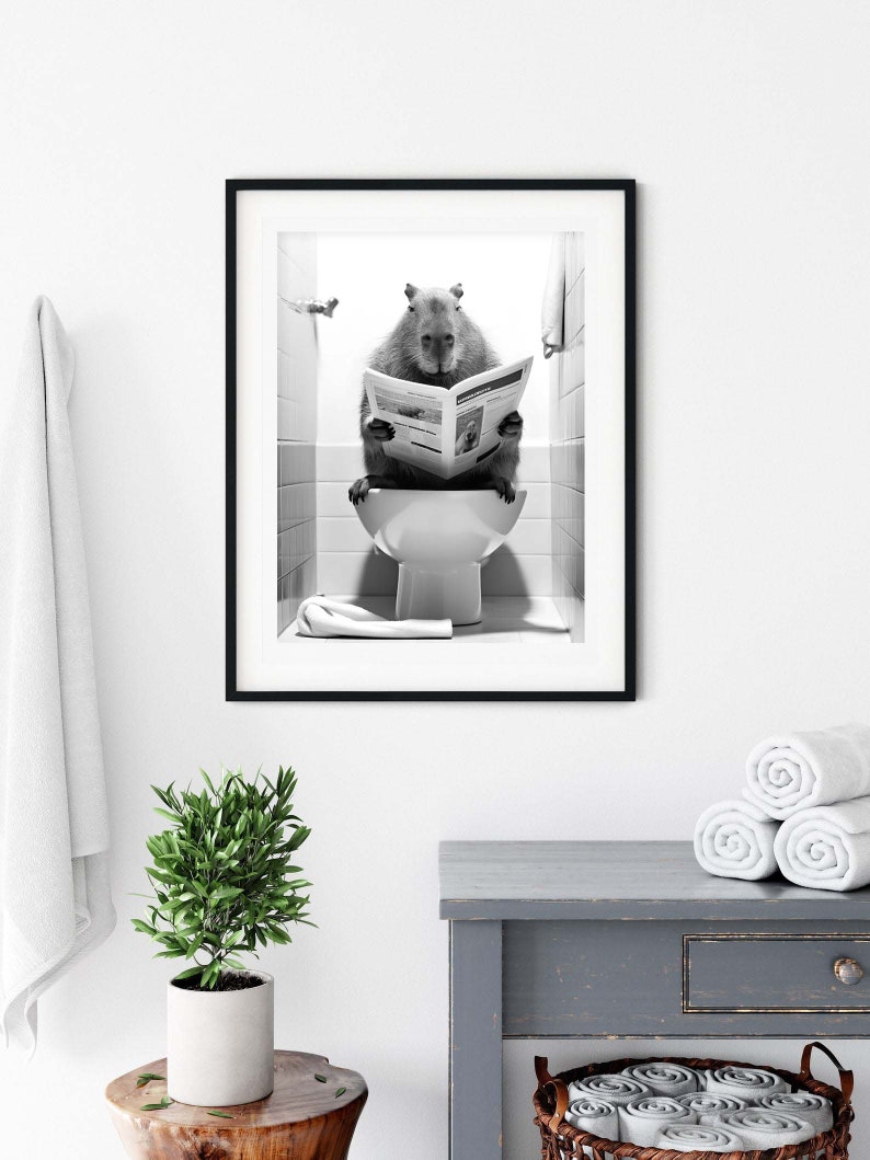 May include: A black and white photograph of a capybara sitting on a toilet and reading a newspaper. The capybara is in a bathroom setting with white tiles and a towel hanging on the wall. The photograph is framed in a black frame.