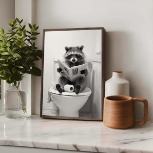 May include: A framed black and white wall art print featuring a raccoon wearing glasses, sitting on a white toilet, and reading a newspaper. The raccoon is holding the open newspaper with both paws, with a roll of toilet paper resting on its lap.