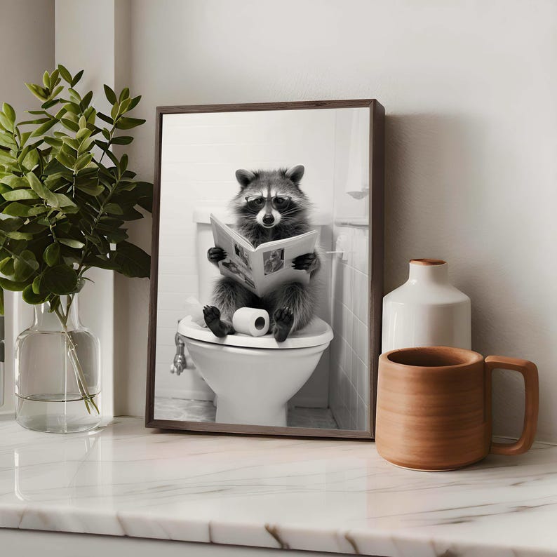 May include: A framed black and white wall art print featuring a raccoon wearing glasses, sitting on a white toilet, and reading a newspaper. The raccoon is holding the open newspaper with both paws, with a roll of toilet paper resting on its lap.