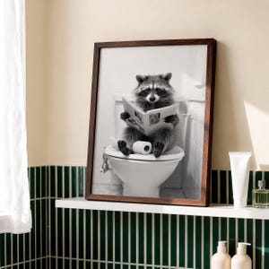 May include: A framed black and white wall art print featuring a raccoon wearing glasses while sitting on a white toilet and reading a newspaper. The raccoon holds the open newspaper with both paws, and a roll of toilet paper sits on the toilet seat.