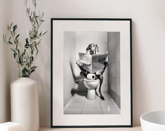 May include: A framed black and white wall art print featuring a dog sitting on a toilet in a tiled bathroom, reading a newspaper. The dog is positioned centrally, holding the open newspaper with its front paws while sitting upright on the white toilet.