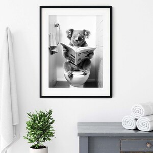 Puede incluir: Una impresi&oacute;n de arte mural enmarcada en blanco y negro que muestra a un koala sentado en un inodoro leyendo un peri&oacute;dico. El koala est&aacute; situado en un ba&ntilde;o con azulejos y una toalla colgada cerca. La obra est&aacute; en un marco negro delgado.