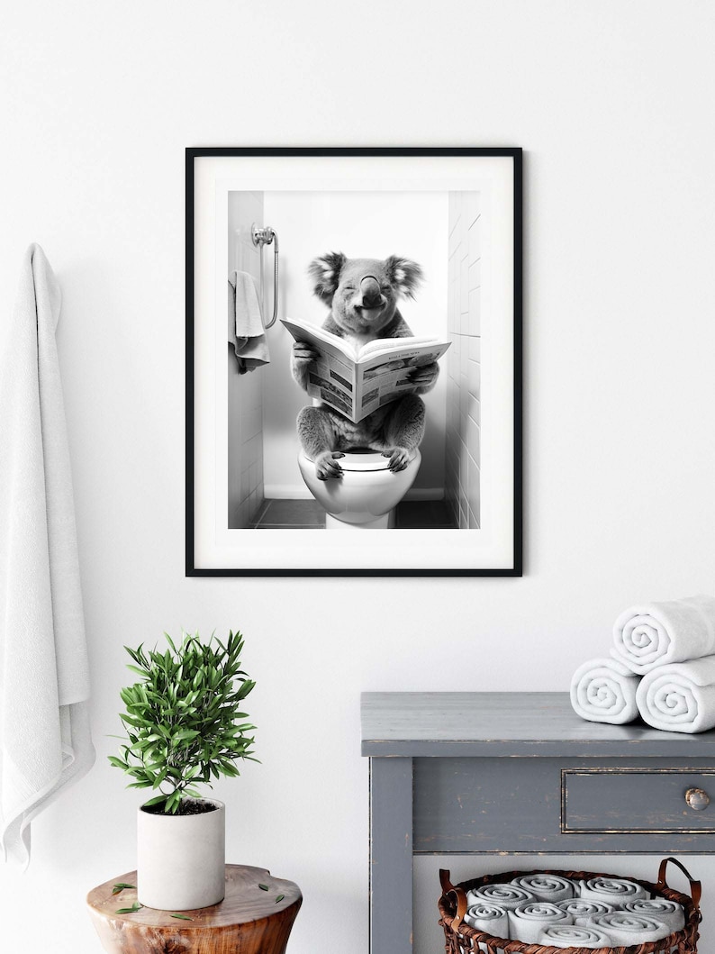 Puede incluir: Una impresi&oacute;n de arte mural enmarcada en blanco y negro que muestra a un koala sentado en un inodoro leyendo un peri&oacute;dico. El koala est&aacute; situado en un ba&ntilde;o con azulejos y una toalla colgada cerca. La obra est&aacute; en un marco negro delgado.