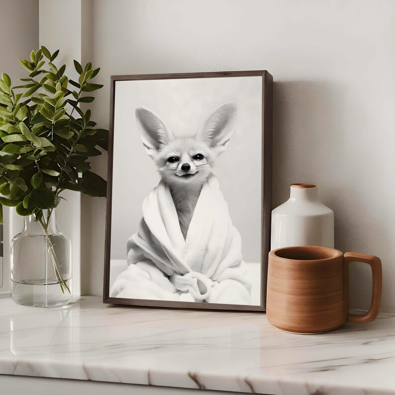 K&ouml;nnte beinhalten: Ein gerahmter Schwarz-Wei&szlig;-Wandkunstdruck, der einen W&uuml;stenfuchs mit gro&szlig;en Ohren zeigt, der in einen wei&szlig;en Bademantel geh&uuml;llt ist. Der Fuchs hat ein dezentes L&auml;cheln und ist im Portr&auml;t zentriert. Der Rahmen ist dunkelbraun und bildet einen Kontrast.