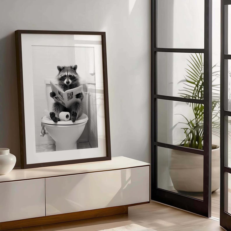 May include: A framed black and white wall art print featuring a raccoon sitting on a white toilet while reading a newspaper. The raccoon is holding the paper with both paws, and a roll of toilet paper sits on the seat next to its feet.