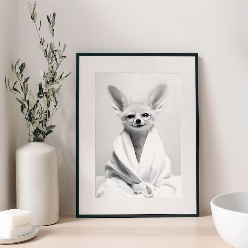 K&ouml;nnte beinhalten: Ein gerahmter Schwarz-Wei&szlig;-Wandkunstdruck, der einen W&uuml;stenfuchs mit gro&szlig;en Ohren zeigt, der in einen wei&szlig;en Pl&uuml;schbademantel geh&uuml;llt ist. Der Fuchs hat ein dezentes L&auml;cheln und ist mittig im Rahmen mit schwarzem Rand und wei&szlig;em Passepartout platziert.