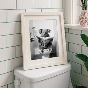 Puede incluir: Una impresi&oacute;n enmarcada en blanco y negro que muestra a un koala sentado en un inodoro blanco mientras lee un peri&oacute;dico abierto. El koala est&aacute; posicionado en el centro de la imagen, sosteniendo el papel con ambas patas, frente a una pared de azulejos.