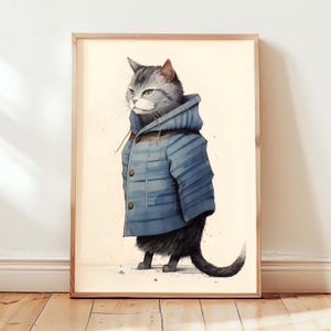 May include: A framed wall art print featuring a grey tabby cat standing upright. The cat is wearing a blue hooded jacket with horizontal stripes and two visible buttons. The illustration is set against a plain, light cream-coloured background.
