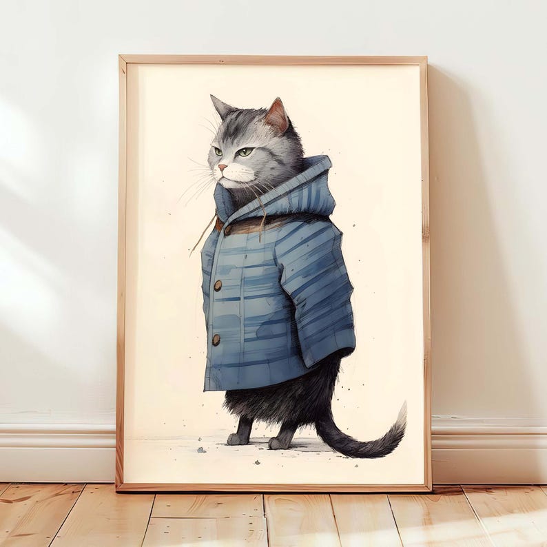 May include: A framed wall art print featuring a grey tabby cat standing upright. The cat is wearing a blue hooded jacket with horizontal stripes and two visible buttons. The illustration is set against a plain, light cream-coloured background.
