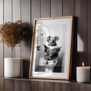 Puede incluir: Una impresi&oacute;n en blanco y negro enmarcada que muestra a un koala sentado en un inodoro mientras lee un peri&oacute;dico. La obra est&aacute; expuesta en un marco de madera clara, colocada en un estante junto a una planta en maceta y una vela blanca.