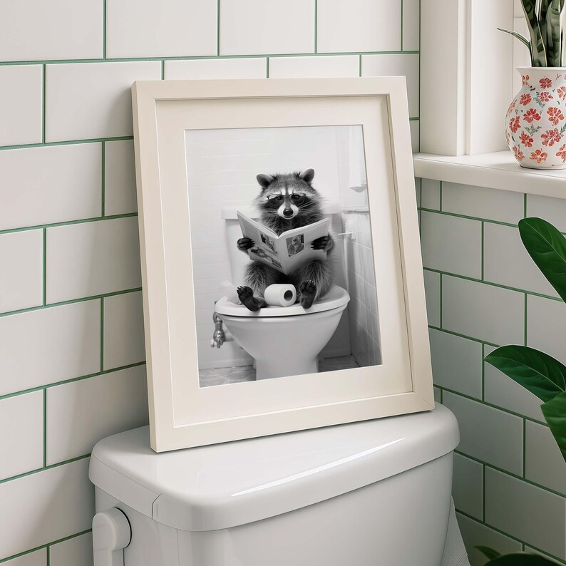 May include: A framed black and white print featuring a raccoon sitting on a white toilet while reading a newspaper. The raccoon is positioned in the center of the image, holding an open newspaper with both paws, with a roll of toilet paper nearby.
