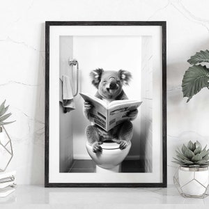 Puede incluir: Una impresi&oacute;n de arte mural enmarcada en blanco y negro que muestra a un koala sentado en un inodoro blanco mientras lee un peri&oacute;dico abierto. El koala tiene una expresi&oacute;n sonriente y est&aacute; en un ba&ntilde;o con azulejos y una toalla en la pared.