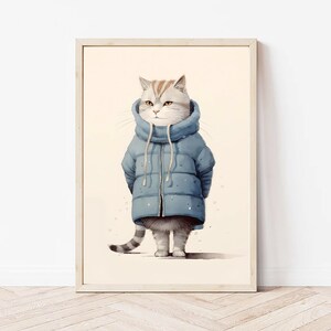 May include: A framed wall art print featuring a light-colored tabby cat wearing a blue puffer jacket with a high collar and drawstrings. The cat has grey and white fur with orange stripes on its head and a striped tail, standing against a cream background.