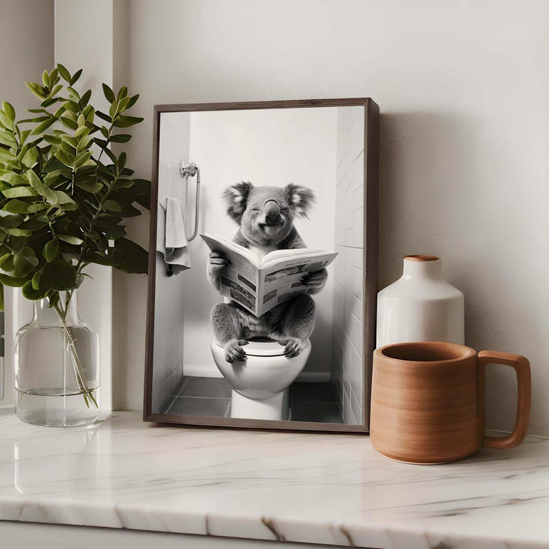 Puede incluir: Una l&aacute;mina de arte mural enmarcada en blanco y negro que muestra a un koala sentado en un inodoro blanco mientras lee un peri&oacute;dico abierto. El koala est&aacute; situado en el centro del marco, con una toalla colgada en un toallero al fondo.