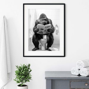 May include: Black and white photograph of a gorilla sitting on a toilet and reading a newspaper. The photo is framed in a black frame and hanging on a white wall.