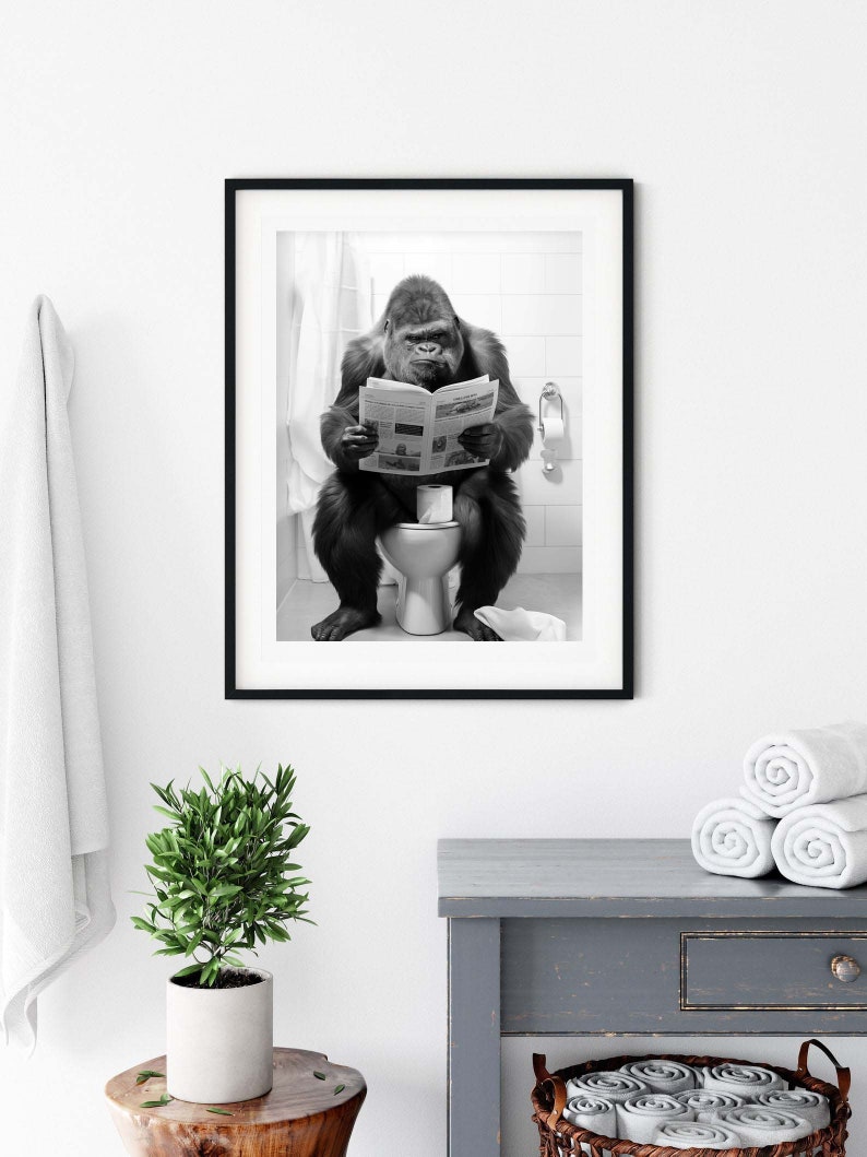 May include: Black and white photograph of a gorilla sitting on a toilet and reading a newspaper. The photo is framed in a black frame and hanging on a white wall.