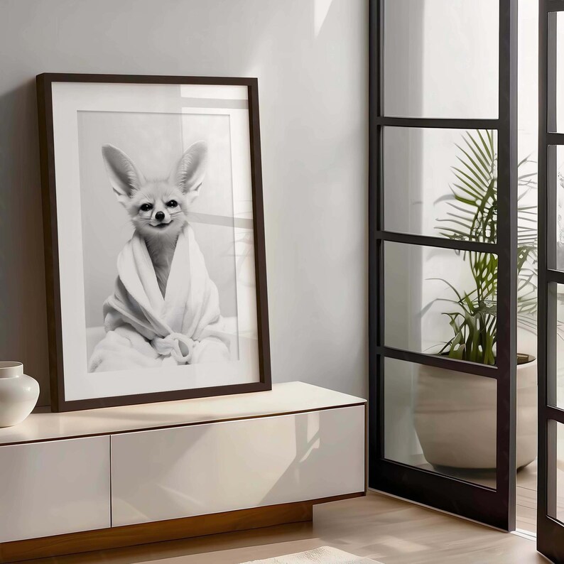 K&ouml;nnte beinhalten: Ein schwarz-wei&szlig;es Wandkunst-Druckbild, das einen W&uuml;stenfuchs in einem wei&szlig;en Bademantel zeigt. Der Fuchs hat gro&szlig;e, spitze Ohren und schaut direkt nach vorne. Das Kunstwerk ist in einem dunkelbraunen rechteckigen Rahmen mit wei&szlig;em Passepartout gefasst.