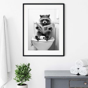 May include: A black and white framed wall art print featuring a raccoon wearing glasses while sitting on a toilet and reading a newspaper. The raccoon is positioned centrally, holding an open newspaper with both paws, with a roll of toilet paper resting on the seat between its feet.