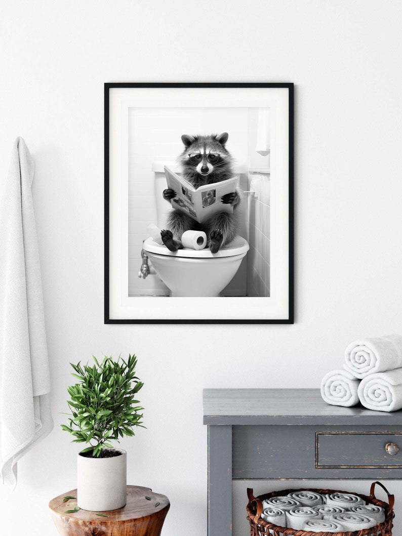 May include: A black and white framed wall art print featuring a raccoon wearing glasses while sitting on a toilet and reading a newspaper. The raccoon is positioned centrally, holding an open newspaper with both paws, with a roll of toilet paper resting on the seat between its feet.