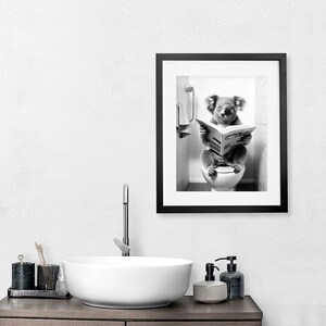 Puede incluir: Una impresi&oacute;n de arte mural enmarcada en blanco y negro que muestra a un koala sentado en un inodoro blanco mientras lee un peri&oacute;dico. El koala est&aacute; en un ba&ntilde;o con una toalla colgada en un soporte cercano. El marco es de color negro.