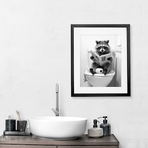 May include: A black and white framed wall art print featuring a raccoon sitting on a white toilet, holding an open newspaper in its paws. The raccoon is positioned centrally on the toilet seat with a roll of toilet paper resting beside its feet.