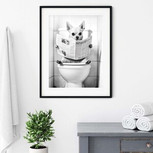 May include: Black and white photo of a small dog sitting on a toilet and reading a newspaper. The dog is wearing a serious expression and is holding the newspaper with its paws. The photo is framed in a black frame.