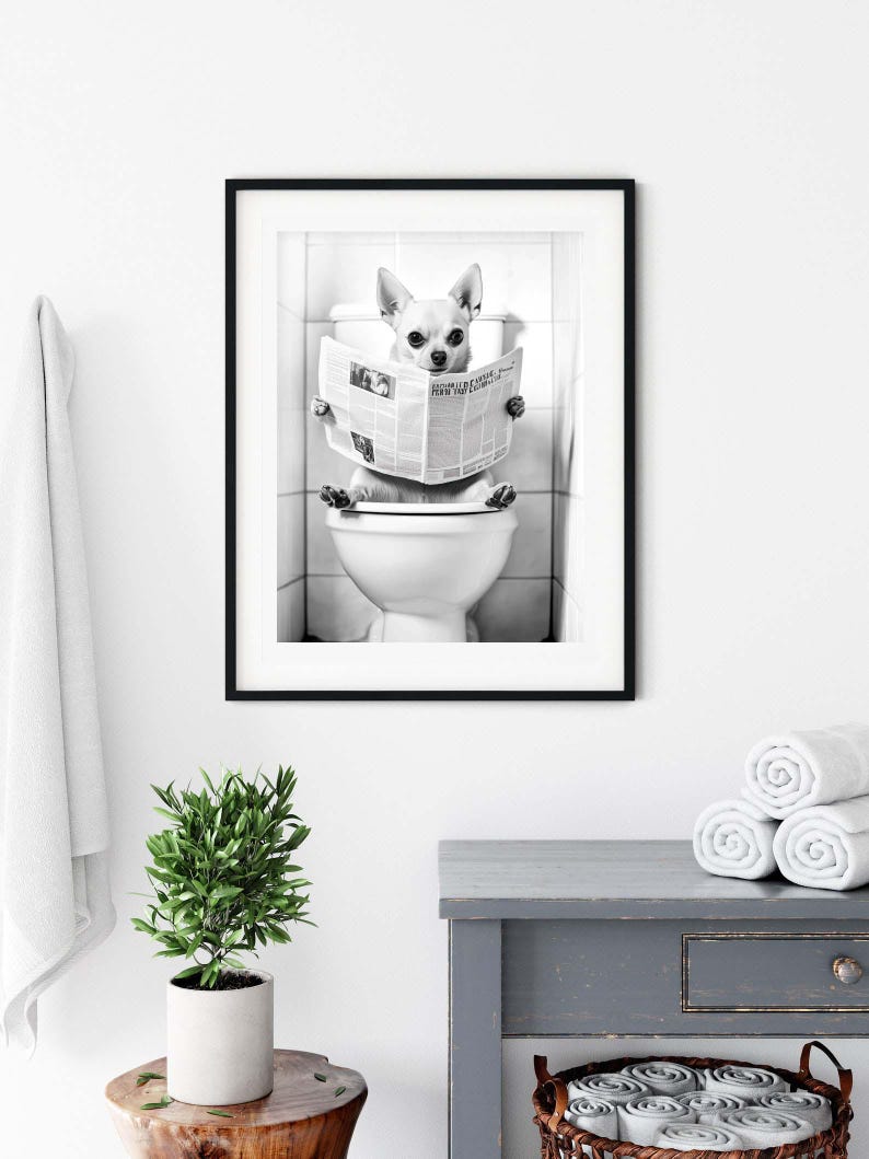 May include: Black and white photo of a small dog sitting on a toilet and reading a newspaper. The dog is wearing a serious expression and is holding the newspaper with its paws. The photo is framed in a black frame.