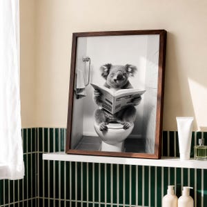 Puede incluir: Una l&aacute;mina de arte mural enmarcada en blanco y negro que muestra a un koala sentado en un inodoro blanco en un ba&ntilde;o, sosteniendo y leyendo un peri&oacute;dico abierto. El koala tiene orejas esponjosas y una expresi&oacute;n sonriente.