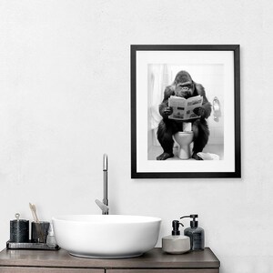 May include: A black and white photograph of a gorilla sitting on a toilet and reading a newspaper. The photo is framed in a black frame and is hanging on a white wall.