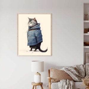 May include: A framed wall art print featuring a grey tabby cat standing in profile. The cat is wearing a blue hooded duffle coat with visible brown toggle buttons. The background of the illustration is a solid, light cream colour.