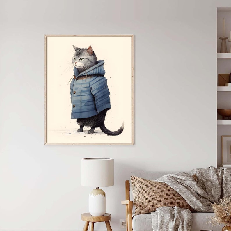 May include: A framed wall art print featuring a grey tabby cat standing in profile. The cat is wearing a blue hooded duffle coat with visible brown toggle buttons. The background of the illustration is a solid, light cream colour.