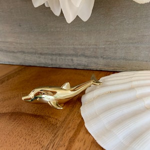 May include: A gold-coloured dolphin figurine on a wooden surface next to a white seashell.
