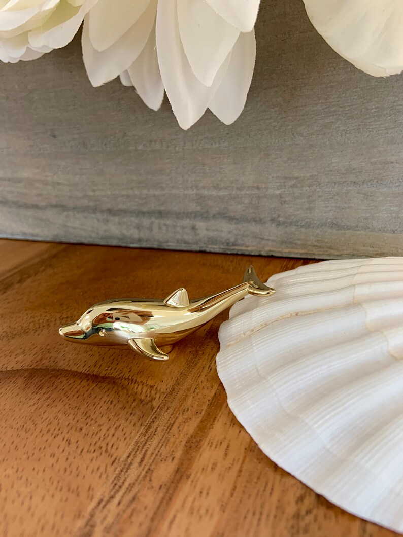 May include: A gold-colored dolphin figurine on a wooden surface next to a white seashell.