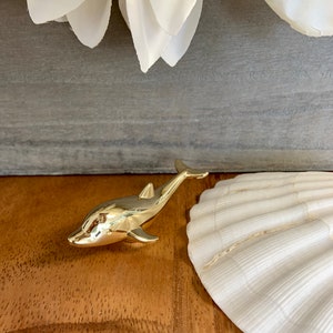 May include: A gold-colored dolphin figurine, with a smooth, shiny finish. The dolphin is positioned on a wooden surface, with a white seashell in the background.