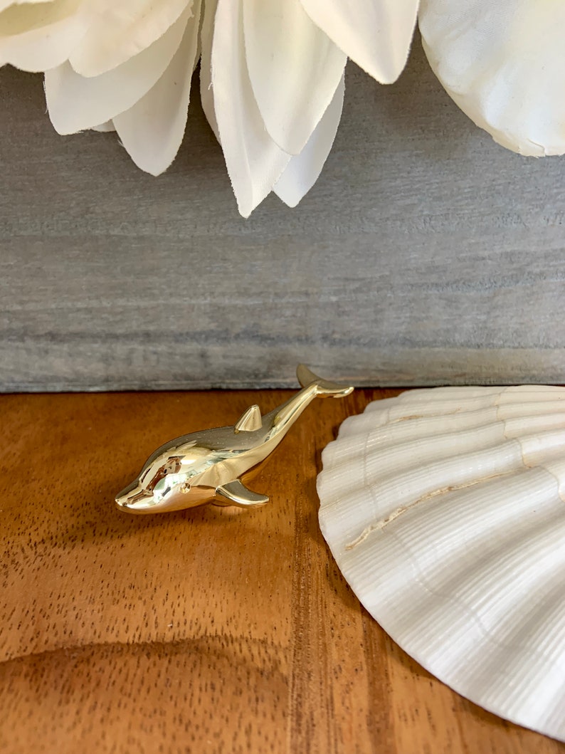 May include: A gold-colored dolphin figurine, with a smooth, shiny finish. The dolphin is positioned on a wooden surface, with a white seashell in the background.