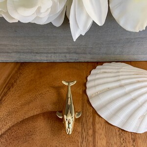 May include: A gold-colored whale figurine on a wooden surface. The whale is facing the camera with its tail up and its fins out. There is a white seashell to the right of the whale.